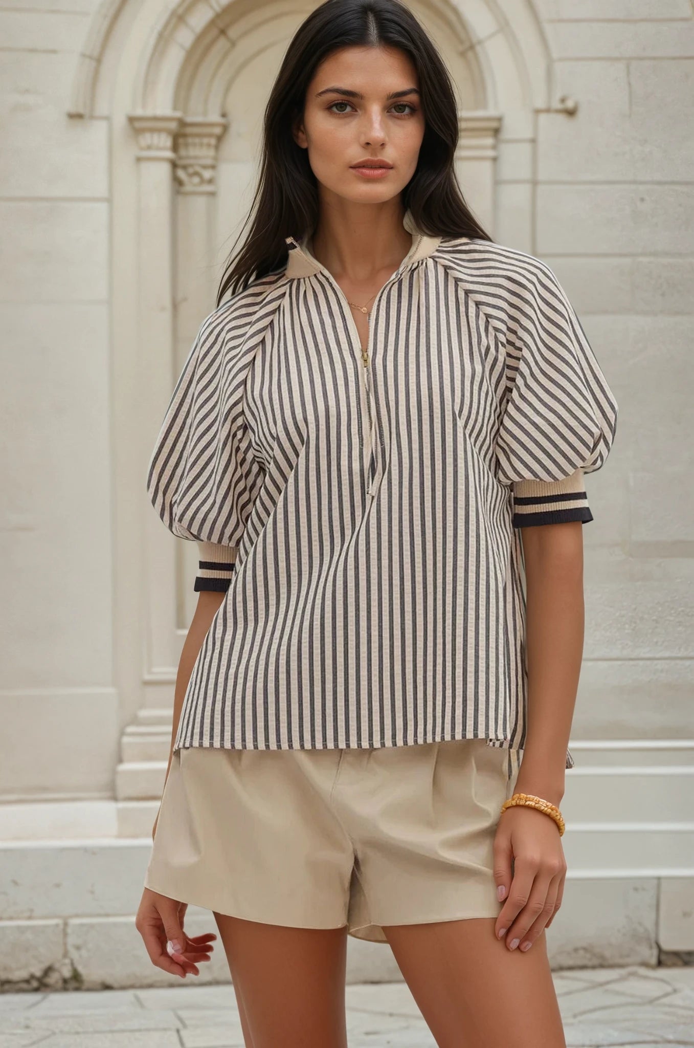 Woman wearing a striped shirt and beige shorts standing in front of a stone building.