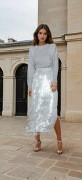 Woman wearing a light gray top and silver sequin skirt standing in front of a building.